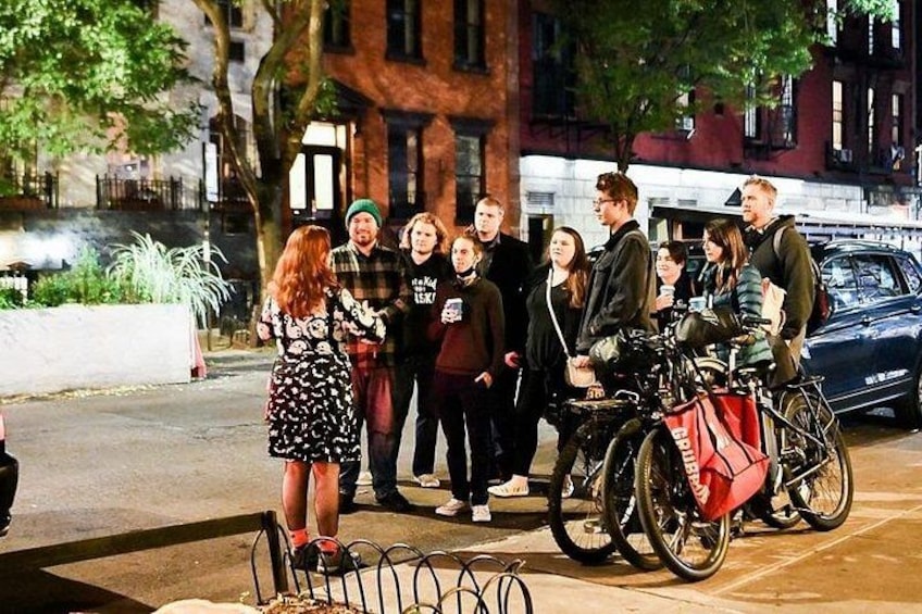 A group of people standing on a sidewalk, some with bicycles, in an urban setting with buildings and trees in the background. 3