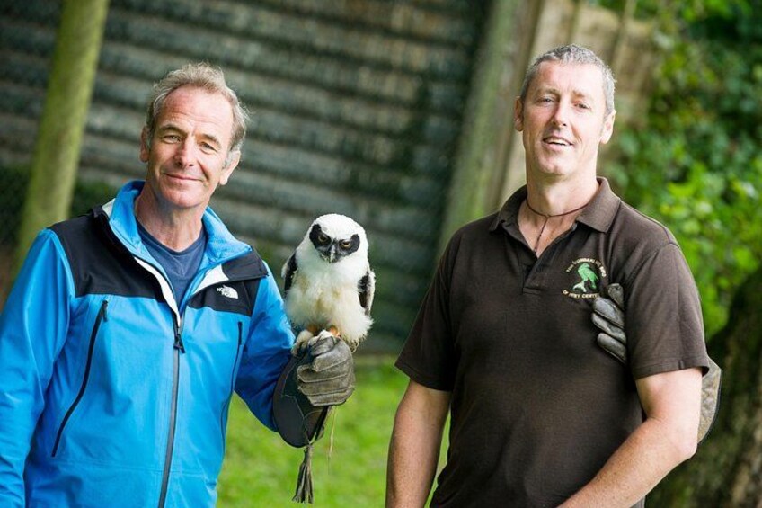 Twee personen houden een vogel vast, buiten met bomen en een houten constructie op de achtergrond. 4