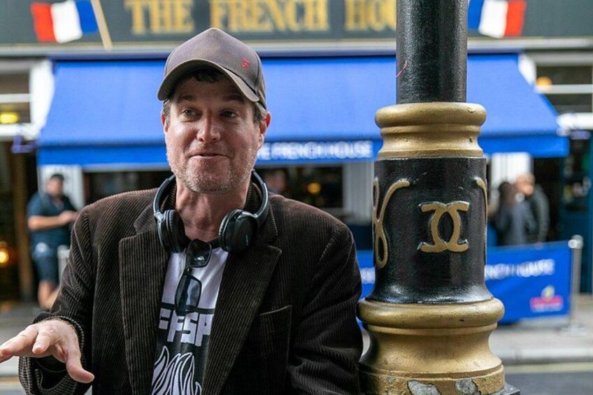 Un hombre con una chaqueta y un gorro marrones, de pie junto a un poste de luz negro y dorado con un emblema 'XX'. 2