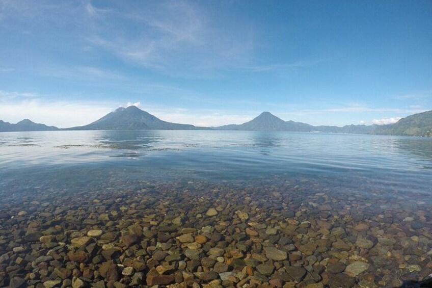 A calm lake with clear water, pebbles at the shore, and mountains in the background. 1