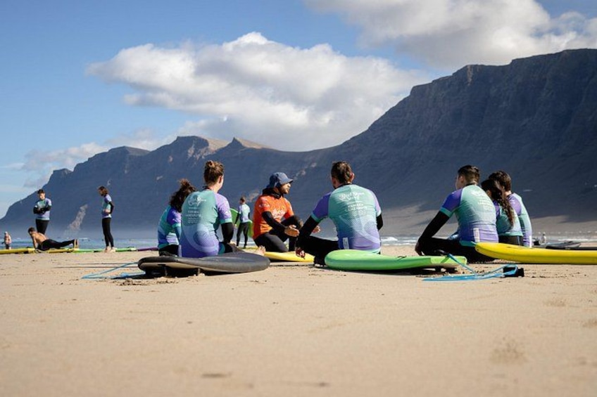 Un gruppo di persone sedute sulla spiaggia con tavole da surf, montagne sullo sfondo e cielo sereno. 5