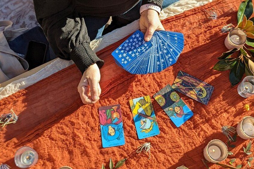 A person performing a tarot card reading with a blue fan and various cards on a sandy surface. 1