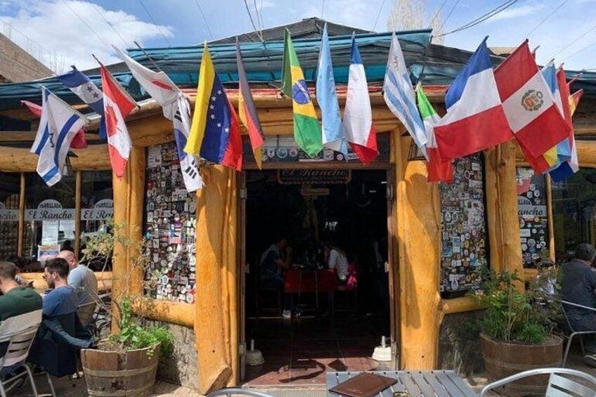A wooden building with multiple flags, including those of Argentina, Brazil, and Chile, and a sign that reads 'El Rancho.' 5