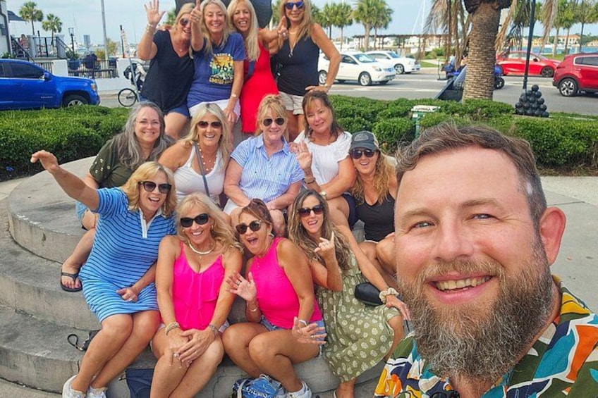 A group of people sitting on a concrete bench outdoors with palm trees and parked cars in the background. 1