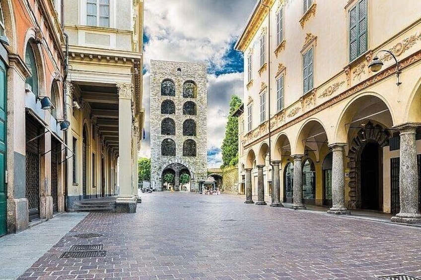 A cobblestone street with historic buildings, an arched entrance, and a central tower. 2