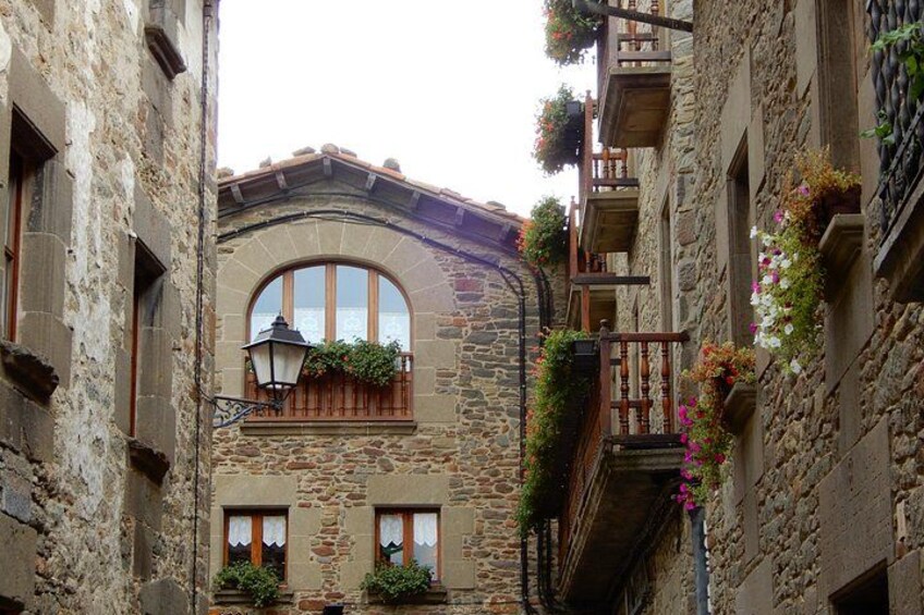 A narrow street with stone buildings, a large arched window, and flower boxes on the balconies. 3