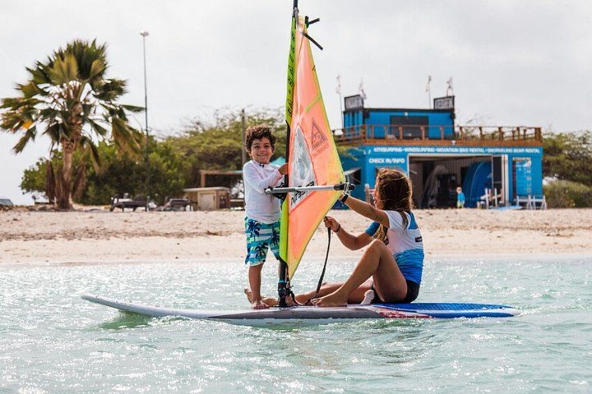 Deux personnes sur une planche de surf avec une voile, une plage, des palmiers et un bâtiment en arrière-plan. 5