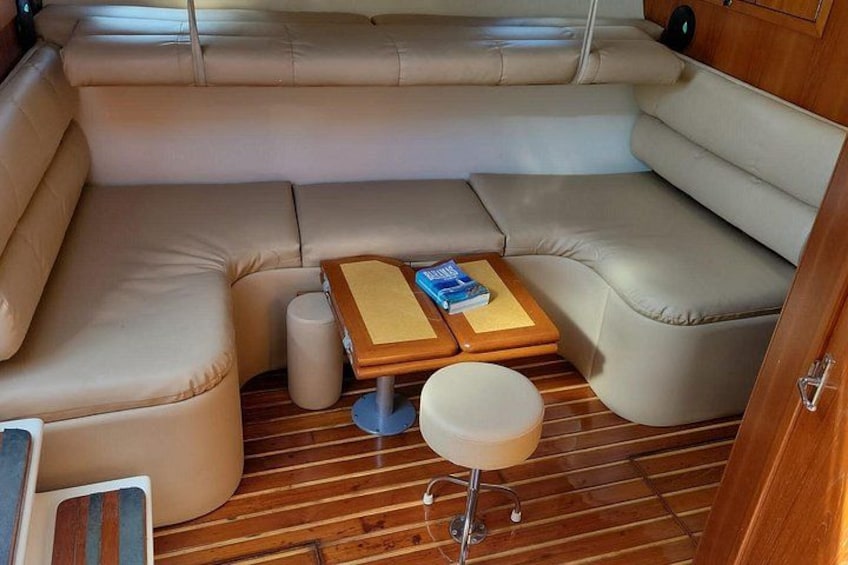 A boat interior with a beige cushioned seating area, a wooden table with a book, and a small stool. 2