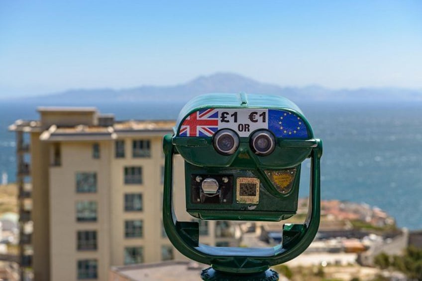Binocoli verdi con una bandiera britannica e un simbolo dell'euro, che si affaccia su una città costiera con edifici e il mare. 5