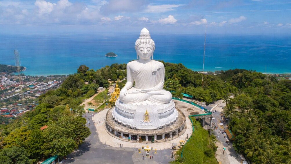A large white Buddha statue surrounded by lush greenery and a coastal view. 1
