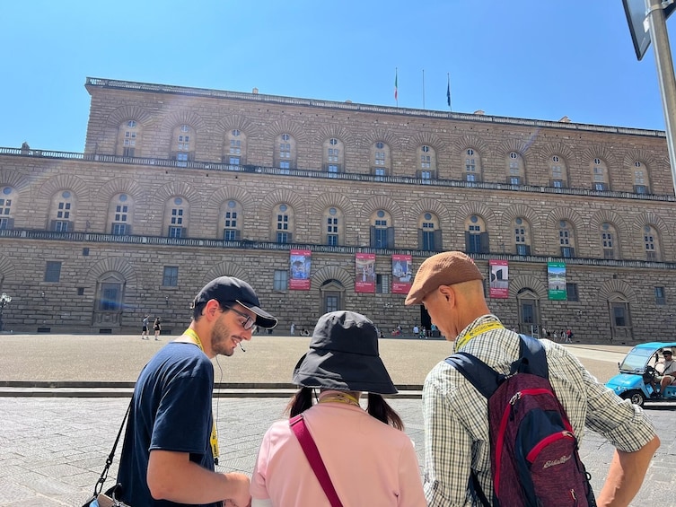 Des touristes se tiennent devant un bâtiment historique doté d’arcades et de fenêtres en arc de cercle, et des drapeaux y sont déployés. 2