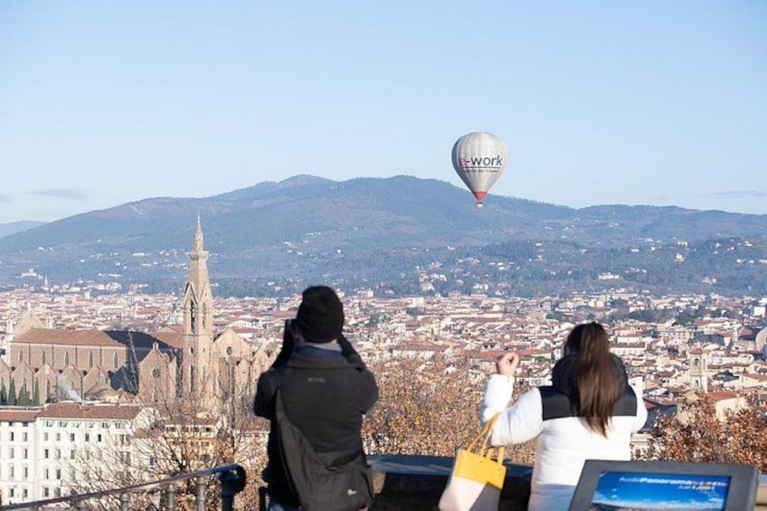 Stadtansicht mit einem markanten Kirchturm, einem Heißluftballon am Himmel und zwei Personen, die zusehen. 5