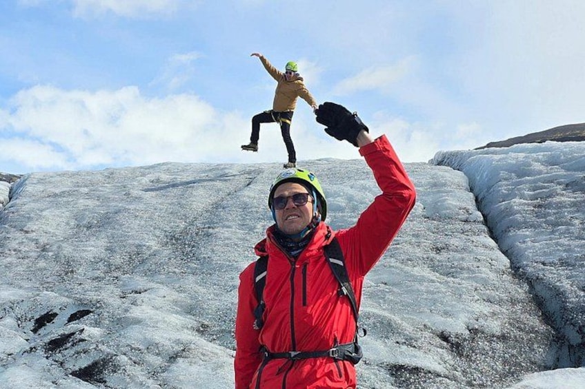 A person in a red jacket and helmet standing on a glacier with another person in a yellow jacket and helmet on a ledge above. 3