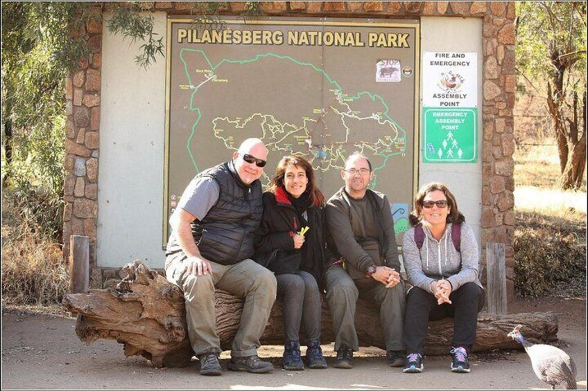 A group of people sitting on a bench in front of a sign for Pilanesberg National Park. 3
