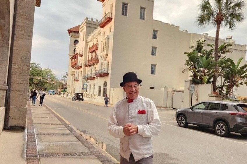 A man in a white chef's jacket standing on a sidewalk with buildings and palm trees in the background. 1