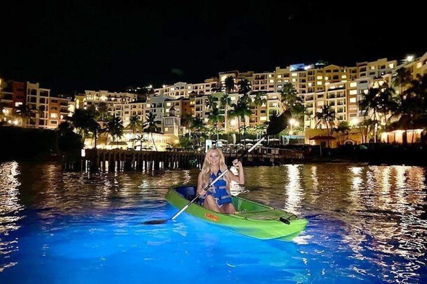 A person in a green kayak on a calm body of water with a brightly lit hotel in the background. 1