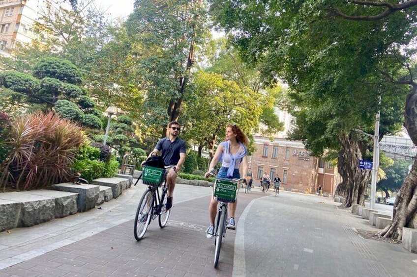 Two people riding bicycles on a paved path surrounded by trees and greenery. 1