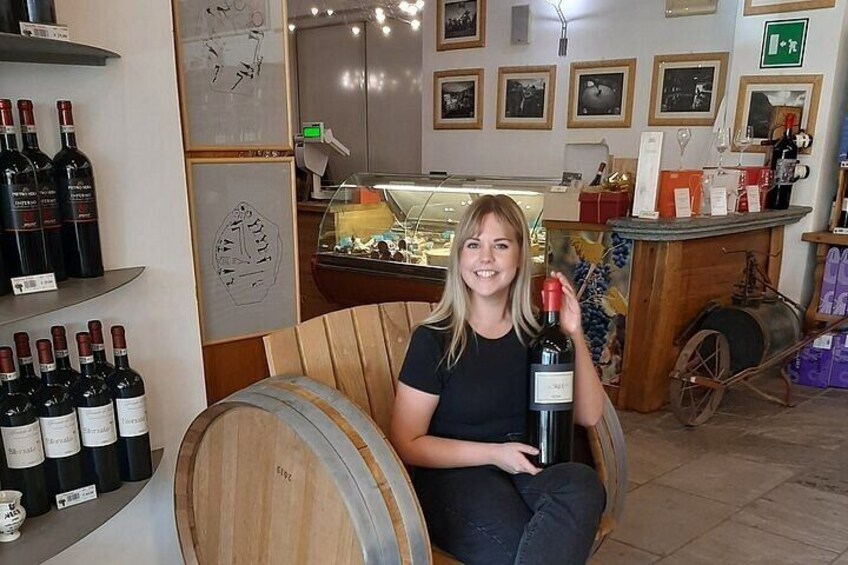 En person sitter på en tunna i en vinbutik, med vinflaskor och en trätunna på golvet. 2