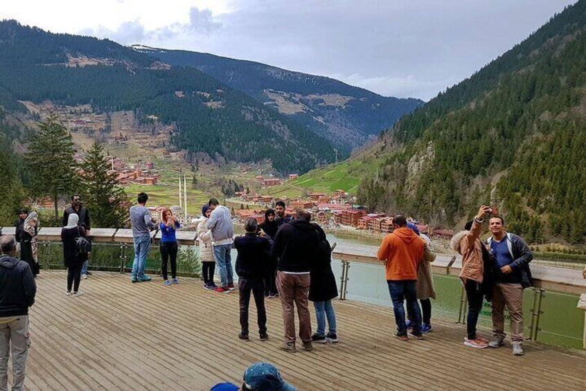 A group of people on a wooden deck overlooking a valley with a river and mountains in the background. 1