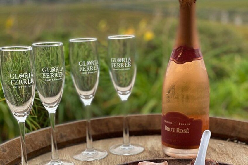 En flaske Brut Rosé vin og fire glas på en træbakke med en vingård i baggrunden. 4