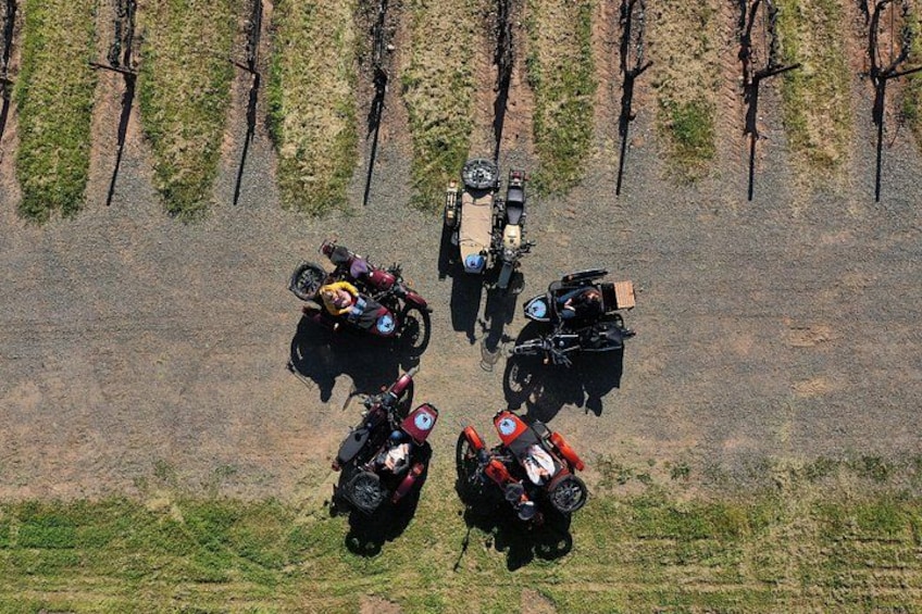Ovenfra ses en gruppe motorcykler parkeret i række på en grusvej, med sparsom vegetation og træer i baggrunden. 2