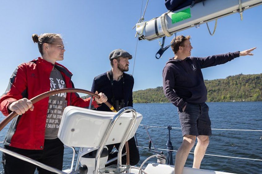 Tres personas en un velero con una costa boscosa al fondo. 1