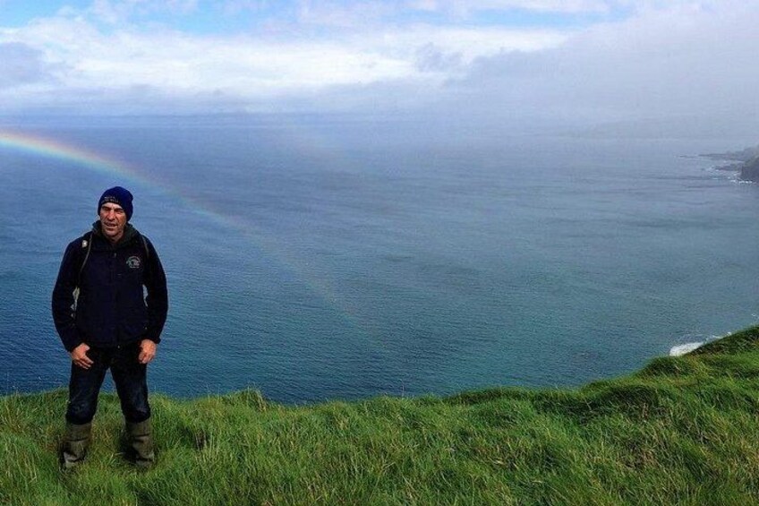 A person standing on a grassy cliff overlooking a vast ocean with a rainbow in the distance. 5