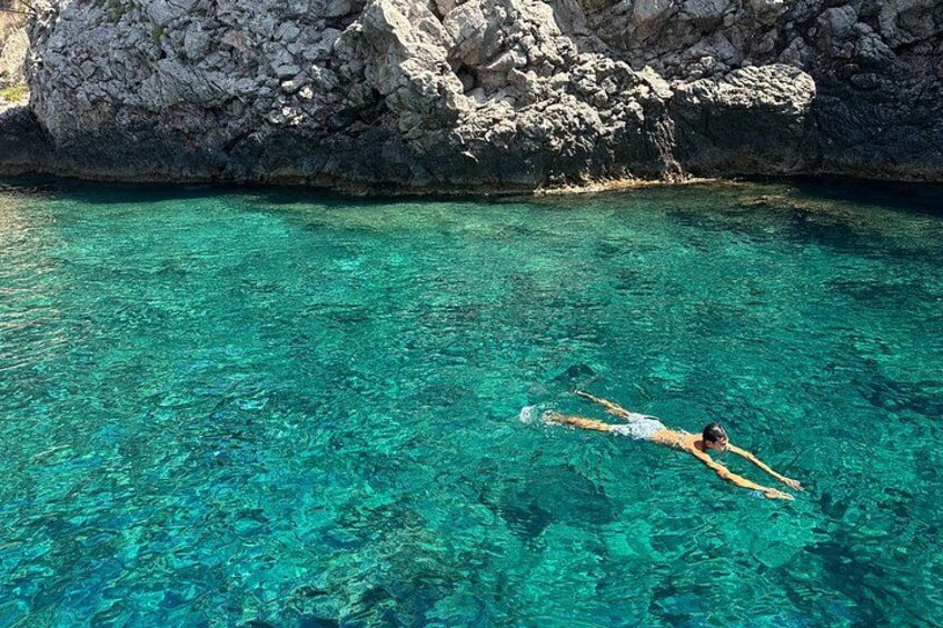 A person swimming in clear turquoise water with rocky formations in the background. 1