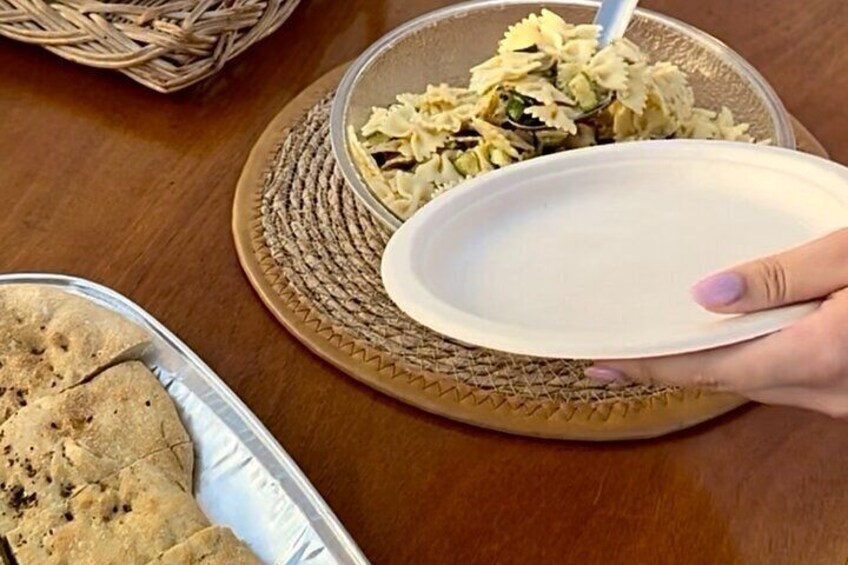 A hand holding a white paper plate over a bowl of pasta. 2