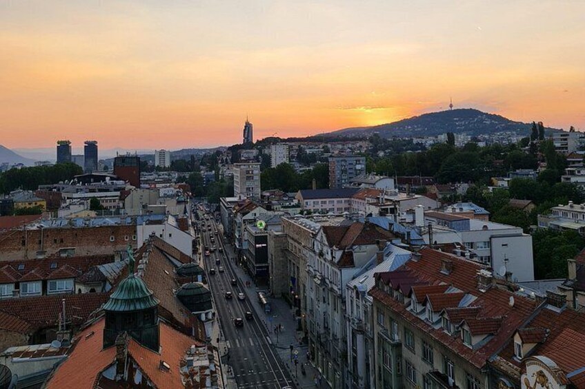 A cityscape at sunset with a prominent tower, a busy street, and a hill in the background. 4