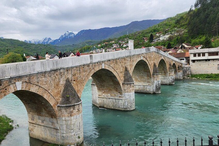 A stone bridge with multiple arches over a river, surrounded by greenery and mountains in the background. 2