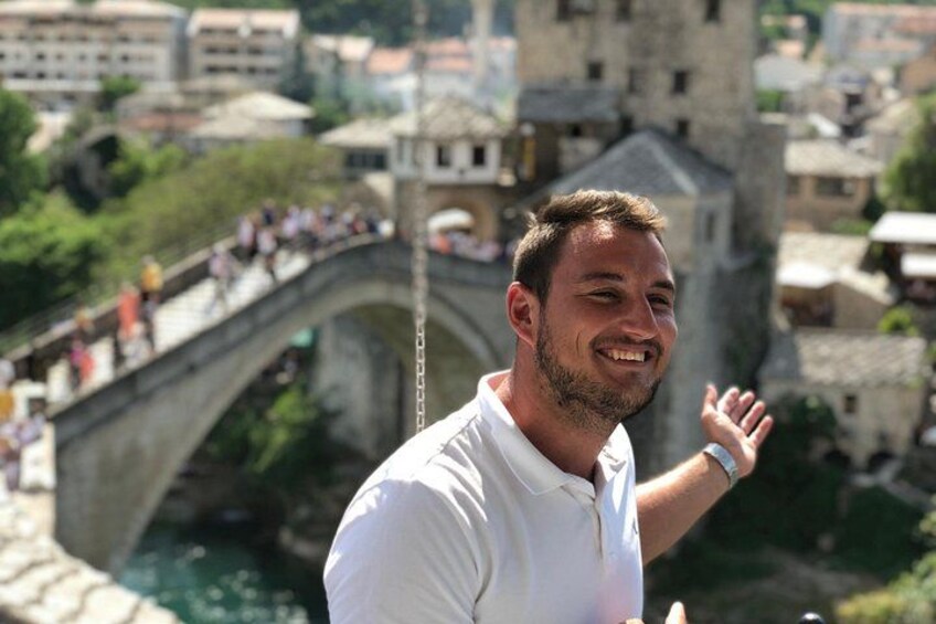 A man in a white shirt standing in front of a stone bridge with a cityscape in the background. 1