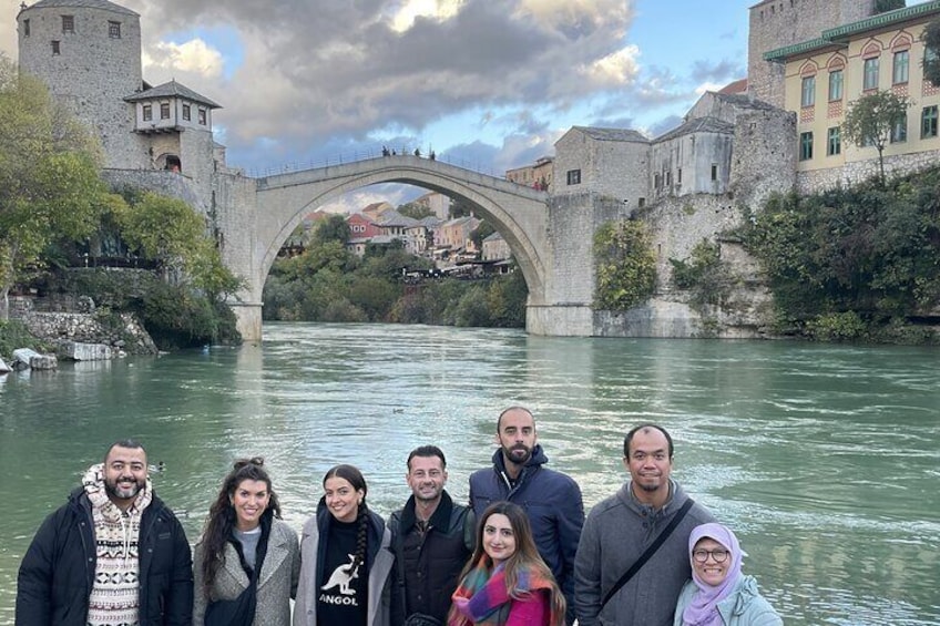 A group of people standing in front of a stone bridge over a river, with historic buildings and greenery in the background. 1