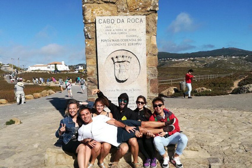 Un groupe de personnes posant devant un monument de pierre orné d’inscriptions, sur fond d’un paysage côtier avec des collines et un ciel dégagé. 2