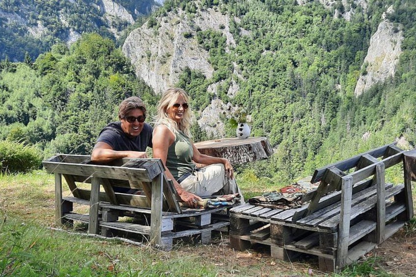 Two people sitting on wooden benches in a mountainous area with greenery and rocky terrain in the background. 2