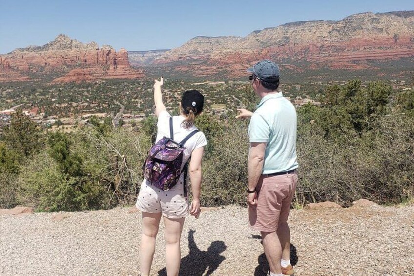 Two people standing on a gravel path overlooking a mountainous landscape with red rock formations and greenery. 2