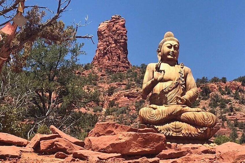 A large Buddha statue in a seated position on a rock formation, surrounded by desert vegetation and a prominent red rock structure in the background. 1