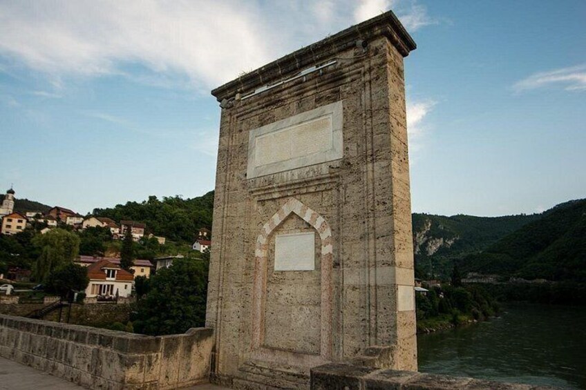 Eine Steinsbrücke mit zwei rechteckigen Fenstern, die auf einen Fluss und eine Stadt mit Häusern und Grünflächen im Hintergrund blickt. 3