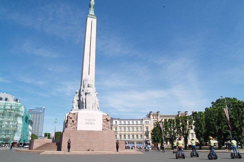 Et højt monument med indskrifter, omgivet af træer og bygninger. 5