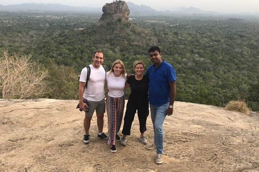 Four people standing on a rocky outcrop with a forested valley and a prominent rock formation in the background. 5