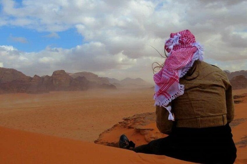 Eine Person sitzt auf einer Sanddüne in einer Wüstenlandschaft, im Hintergrund sind Berge zu sehen. 3