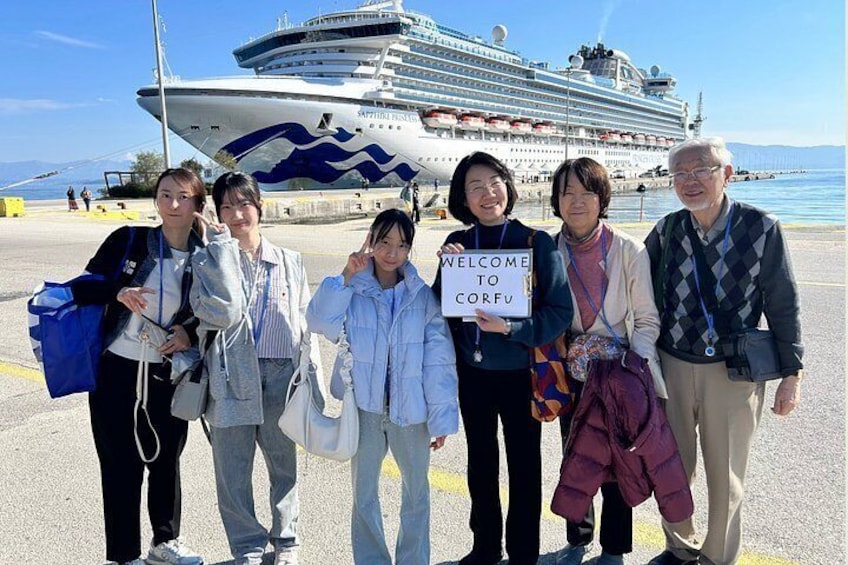 Un groupe de personnes se tient devant un navire de croisière, une pancarte indiquant : « BIENVENUE À CORFOU ». 1