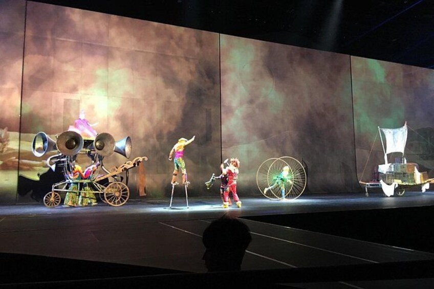 A stage performance with performers in costumes and props, including a large wheel and a character on stilts. 2