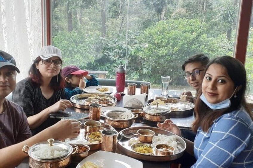Un grupo de personas cenando en una mesa con varios platos y vistas a follaje exterior. 4