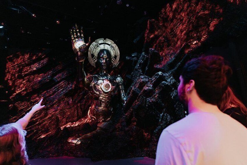 A metallic statue with a raised hand, set against a rocky background. 2