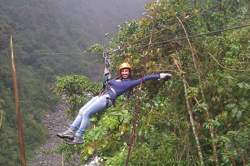 En person åker på en zipline genom ett skogsområde med tät vegetation, och en flod syns i bakgrunden. 1
