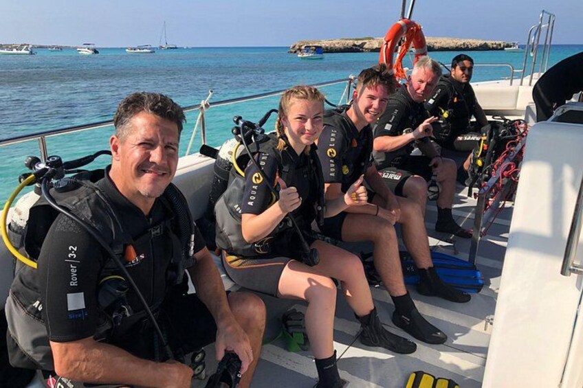 Un groupe de personnes se trouve sur un bateau, dont certains portent l’équipement de plongée sous-marine. On aperçoit une mer bleue et limpide ainsi que d’autres bateaux à l’arrière-plan. 1