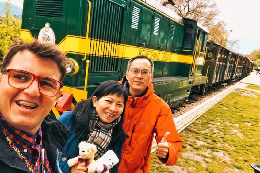 A green and yellow train on tracks, a person in an orange jacket, and two individuals holding teddy bears. 5