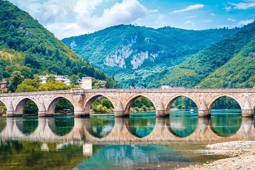 A stone bridge with multiple arches over a river, surrounded by lush green hills and a clear sky. 1