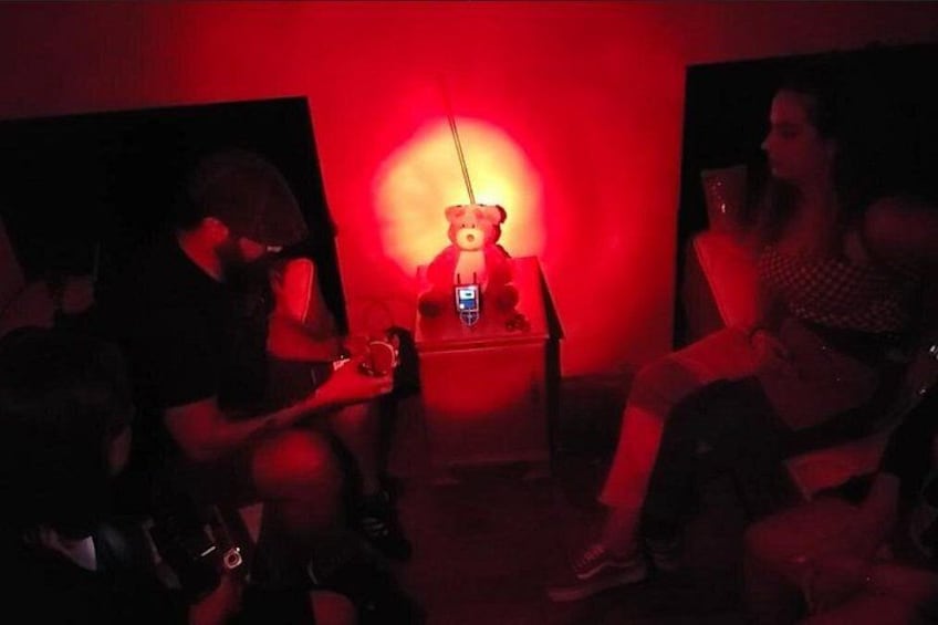 Ein schwach beleuchteter Raum mit rotem Licht, einem Teddybären und zwei Personen, die auf Stühlen sitzen. 4